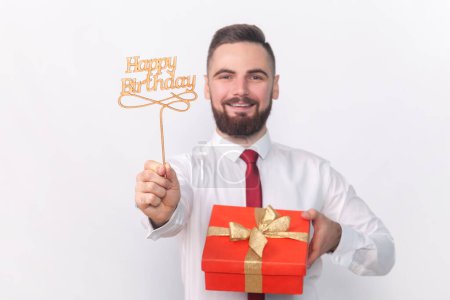 Photo for Portrait of smiling satisfied handsome man in white shirt holding party props with happy birthday inscription and red present box. Indoor studio shot isolated on gray background. - Royalty Free Image