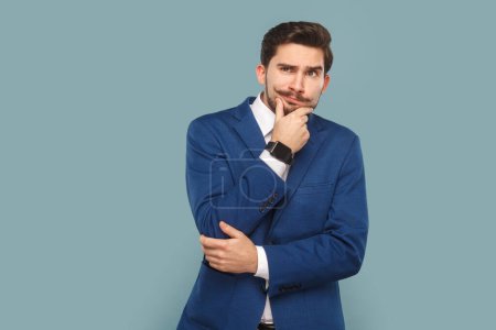 Foto de Pensive thoughtful handsome man standing and holding his chin, thinking about future, looking away, wearing white shirt and jacket. Indoor studio shot isolated on light blue background. - Imagen libre de derechos