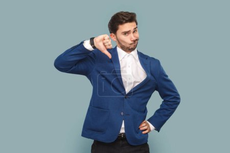 Photo for Portrait of dissatisfied attractive man with mustache with frowning face, showing thumb up, negative feedback, wearing white shirt and jacket. Indoor studio shot isolated on light blue background. - Royalty Free Image
