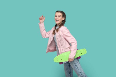 Téléchargez les photos : Portrait of extremely happy funny teenager girl with braids wearing pink jacket standing with raised arm, holding skateboard in hands. Indoor studio shot isolated on green background. - en image libre de droit