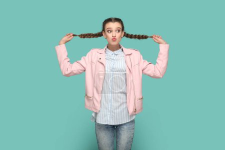 Téléchargez les photos : Portrait of funny positive flirting teenager girl with braids wearing pink jacket pulling her pigtails, sending air kissing, posing with pot lips. Indoor studio shot isolated on green background. - en image libre de droit