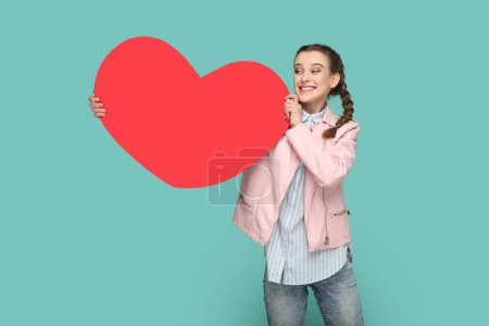 Foto de Portrait of extremely happy cheerful teenager girl with braids wearing pink jacket, showing big red heart, looking away with toothy smile. Indoor studio shot isolated on green background. - Imagen libre de derechos