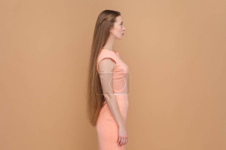 Foto de Side view portrait of serious strict woman with long hair looking ahead with bossy expression, being in bad mood, wearing elegant dress. Indoor studio shot isolated on brown background. - Imagen libre de derechos