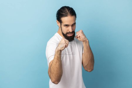 Foto de Portrait of aggressive man with beard wearing white T-shirt holding clenched fists up ready to boxing, martial art trainer, self defense. Indoor studio shot isolated on blue background. - Imagen libre de derechos