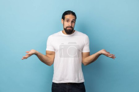 Photo for So what to do in this situation I dont know. Hesitant clueless man with beard wearing white T-shirt shrugging shoulders, spreads palms feels unaware. Indoor studio shot isolated on blue background. - Royalty Free Image