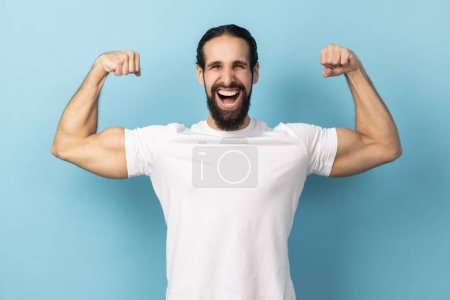 Téléchargez les photos : Portrait d'homme indépendant bodybuilder confiance en soi portant T-shirt blanc montrant biceps levant les bras forts, fier de la construction musculaire, le leadership. Studio intérieur tourné isolé sur fond bleu. - en image libre de droit