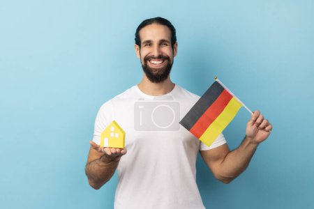 Téléchargez les photos : Portrait of happy handsome man with beard wearing white T-shirt holding german flag and paper house, dreaming to buy apartment in Germany. Indoor studio shot isolated on blue background. - en image libre de droit