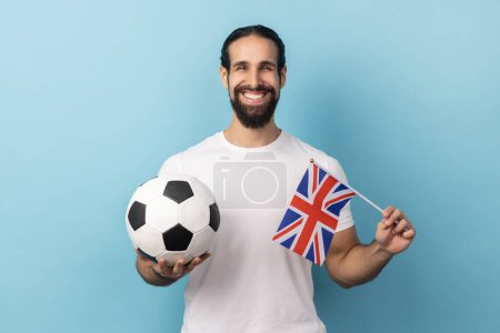 Téléchargez les photos : Portrait d'homme satisfait avec barbe portant un T-shirt blanc tenant le drapeau de l'Angleterre et le ballon noir et blanc de football, ligue de football unie. Studio intérieur tourné isolé sur fond bleu. - en image libre de droit
