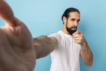 Foto de Portrait of attractive handsome man with beard wearing white T-shirt looking at camera and pointing, POV, point of view of photo, choosing you. Indoor studio shot isolated on blue background. - Imagen libre de derechos