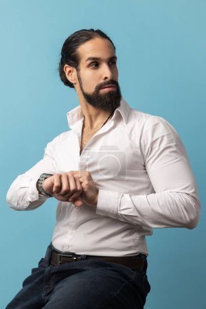 Téléchargez les photos : Handsome confident man with beard wearing white shirt sitting and touching his wristwatch, looking away, being harry to important event. Indoor studio shot isolated on blue background. - en image libre de droit