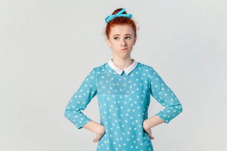 Foto de Portrait of confused puzzled ginger woman standing with hands on hips, looking away, thinking, keeps hands on hips, wearing blue dress. Indoor studio shot isolated on gray background. - Imagen libre de derechos