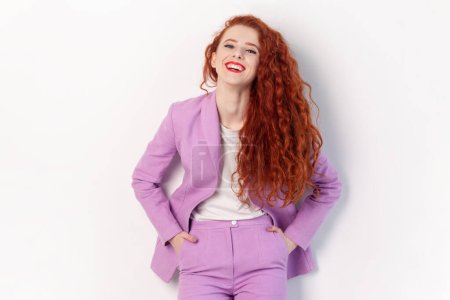 Téléchargez les photos : Portrait d'une heureuse femme aux cheveux roux, heureuse et positive, aux cheveux ondulés, les mains dans les poches, regardant la caméra et riant, portant un costume lilas. Studio intérieur tourné isolé sur fond gris - en image libre de droit