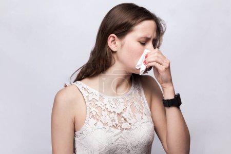 Téléchargez les photos : Portrait of unhealthy attractive young adult woman wearing white dress catching cold, having flu symptoms and runny nose, sneezing. Indoor studio shot isolated on gray background. - en image libre de droit
