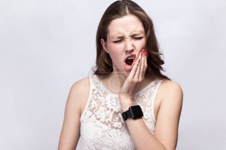 Téléchargez les photos : Portrait of stressed woman wearing white dress standing with frowning face and closed eyes, feeling terrible toothache, needs to visit dentist. Indoor studio shot isolated on gray background. - en image libre de droit
