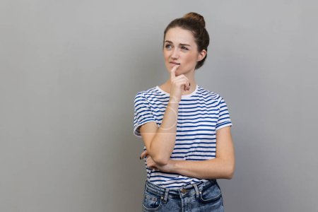 Photo for Portrait of woman wearing striped T-shirt pondering serious issues, looking with uncertain hesitant expression, making difficult choice. Indoor studio shot isolated on gray background. - Royalty Free Image