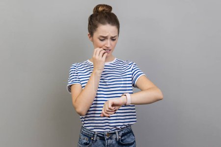 Photo for I need more time. Portrait of nervous woman wearing striped T-shirt standing and looking at her smart watch and want more time, biting fingernails. Indoor studio shot isolated on gray background. - Royalty Free Image