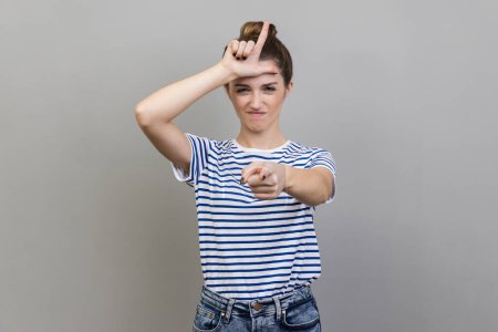 Photo for Portrait of woman wearing striped T-shirt expressing disrespect, showing L finger sign to camera, loser gesture, accusing for failure. Indoor studio shot isolated on gray background. - Royalty Free Image