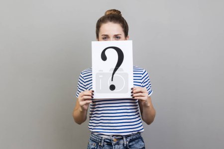 Photo for Portrait of woman wearing striped T-shirt holding white billboard with question mark on it, covering half of face with, looking at camera. Indoor studio shot isolated on gray background. - Royalty Free Image