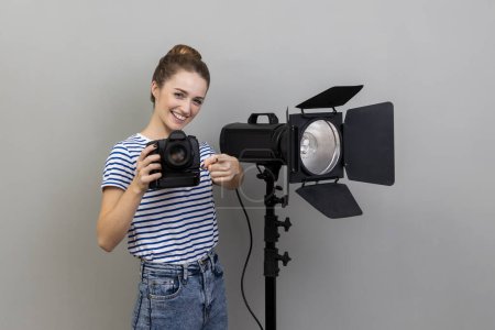 Téléchargez les photos : Portrait of smiling satisfied woman photographer photographing, holding photo camera and using illumination equipment, pointing to camera. Indoor studio shot isolated on gray background. - en image libre de droit