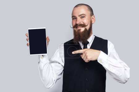 Téléchargez les photos : Portrait of smiling satisfied man with beard and mustache standing pointing at tablet with black blank screen, copy space for advertisement. Indoor studio shot isolated on gray background. - en image libre de droit
