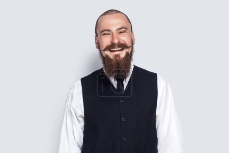 Téléchargez les photos : Portrait of excited attractive young adult man with beard and mustache standing and laughing, hearing funny jokes, looking at camera. Indoor studio shot isolated on gray background. - en image libre de droit