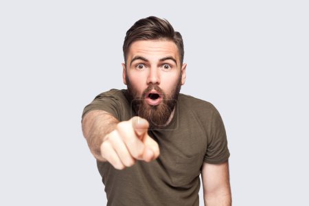 Téléchargez les photos : Surprised wondered bearded man wearing dark green T-shirt pointing fingers looking at camera with open mouth and big eyes, shocked with choice. Indoor studio shot isolated on light gray background. - en image libre de droit