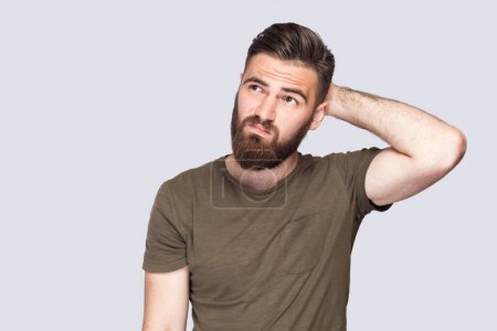 Téléchargez les photos : Portrait of pensive bearded man wearing dark green T-shirt touching head and thinking about important question, making hard decision. Indoor studio shot isolated on light gray background. - en image libre de droit