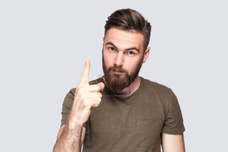 Photo for Portrait of serious attractive bearded man wearing dark green T-shirt standing with admonishing gesture, scolding for mistake and warning. Indoor studio shot isolated on light gray background. - Royalty Free Image