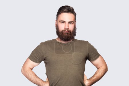 Photo for Portrait of bearded man wearing dark green T-shirt looking cross-eyed with stupid facial expression, fooling around, keeps hands on hips. Indoor studio shot isolated on light gray background. - Royalty Free Image