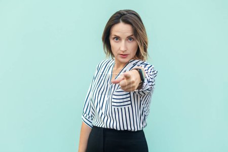 Téléchargez les photos : Portrait of angry serious middle aged woman wearing striped shirt standing pointing to camera, choosing you, looking at bossy expression. Indoor studio shot isolated on light blue background. - en image libre de droit