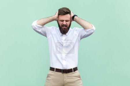 Photo for Portrait of stressed depressed bearded businessman wearing white shirt standing with hands on head and screaming, suffering terrible headache. Indoor studio shot isolated on light green background. - Royalty Free Image