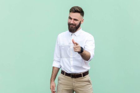 Photo for Hey, I choose you. Portrait of attractive flirting bearded businessman wearing white shirt pointing finger and winking at camera, choosing you. Indoor studio shot isolated on light green background. - Royalty Free Image