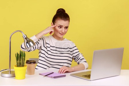 Photo for Woman office worker showing stupid gesture with finger to laptop screen with displeased tired expression, sitting at workplace. Indoor studio studio shot isolated on yellow background. - Royalty Free Image