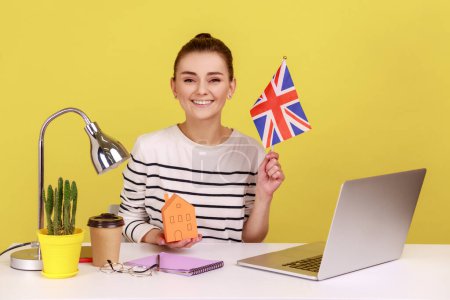 Mujer feliz optimista mostrando la bandera de Gran Bretaña, lugar de trabajo sentado con la casa de papel, satisfecho con la compra de alojamiento. Estudio de interior plano aislado sobre fondo amarillo.