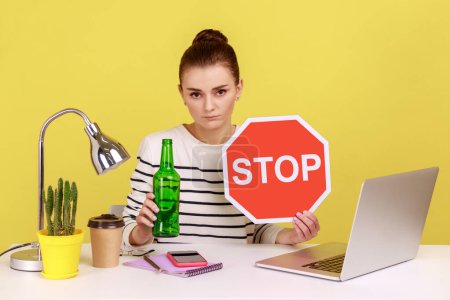 Photo for Portrait of anxious beautiful woman showing alcoholic beverage beer bottle and stop sign, sitting on workplace with laptop. Indoor studio studio shot isolated on yellow background. - Royalty Free Image