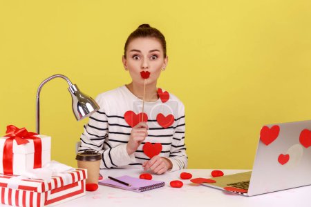 Foto de Positive funny woman office manager sitting on workplace covered with heart sticks, holding party props, covering her mouth with paper lips. Indoor studio studio shot isolated on yellow background. - Imagen libre de derechos
