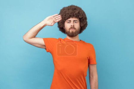 Téléchargez les photos : Portrait of man with Afro hairstyle wearing orange T-shirt saluting with respect as if soldier waiting order from commander, obeying discipline. Indoor studio shot isolated on blue background. - en image libre de droit