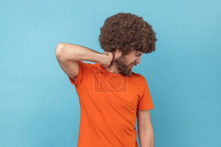 Foto de Portrait of man with Afro hairstyle in T-shirt touching neck, feeling acute pain moving and turning head, suffering spine problems, osteochondrosis. Indoor studio shot isolated on blue background. - Imagen libre de derechos