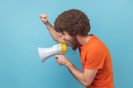 Photo for Side view of serious man with Afro hairstyle wearing orange T-shirt raised hands and holding megaphone, screaming in loud speaker, protesting. Indoor studio shot isolated on blue background. - Royalty Free Image