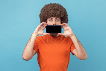Photo for Portrait of funny positive man with Afro hairstyle in orange T-shirt covering mouth with smart phone with empty display, looking at camera with big eyes. Indoor studio shot isolated on blue background - Royalty Free Image
