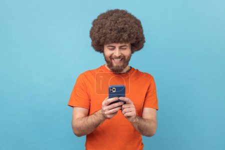 Téléchargez les photos : Portrait d'un blogueur homme positif avec une coiffure afro portant un T-shirt orange debout, utilisant un smartphone et regardant une vidéo avec un visage souriant heureux. Studio intérieur tourné isolé sur fond bleu. - en image libre de droit