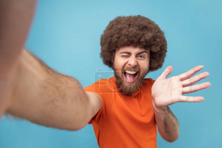 Téléchargez les photos : Portrait of excited man with Afro hairstyle wearing orange T-shirt having livestream, talking with followers, waving hand, greeting, POV. Indoor studio shot isolated on blue background. - en image libre de droit