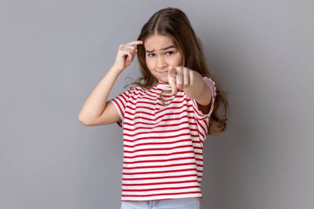 Téléchargez les photos : You're idiot with insane crazy plan. Little girl wearing striped T-shirt showing stupid gesture and pointing to camera, accusing of dumb idea. Indoor studio shot isolated on gray background. - en image libre de droit