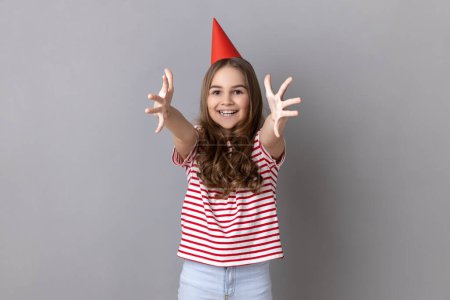 Foto de Portrait of pretty little girl wearing striped T-shirt and party cone on head giving free hugs with outstretched hands, welcoming inviting to embrace. Indoor studio shot isolated on gray background. - Imagen libre de derechos