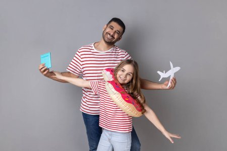 Foto de Portrait of excited smiling father and daughter in striped T-shirts holding passport and rubber ring, journey abroad, vacation to the sea. Indoor studio shot isolated on gray background. - Imagen libre de derechos