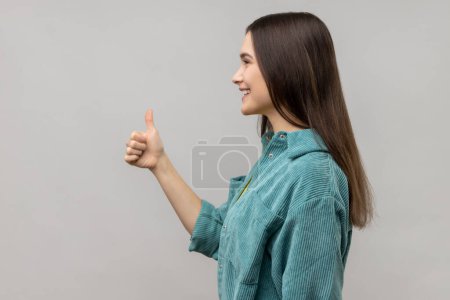 Téléchargez les photos : Side view of woman looking ahead with smile and showing thumb up, approval sign, satisfied with service, good feedback, wearing casual style jacket. Indoor studio shot isolated on gray background. - en image libre de droit