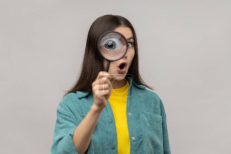 Foto de Surprised woman standing, holding magnifying glass and looking at camera with big zoom eye, having amazed expression, wearing casual style jacket. Indoor studio shot isolated on gray background. - Imagen libre de derechos