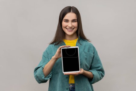 Foto de Portrait of positive delighted woman holding showing tablet, looking at camera with smile and happiness, wearing casual style jacket. Indoor studio shot isolated on gray background. - Imagen libre de derechos