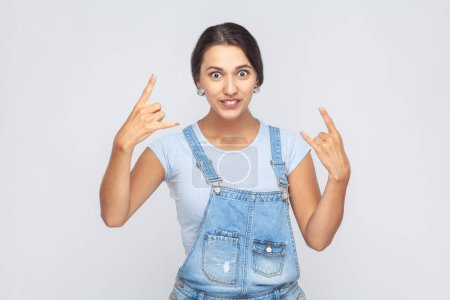 Photo for Portrait of beautiful young adult woman wearing denim overalls showing rock and roll hand sign, looking crazy with devil horn gesture. Indoor studio shot isolated on gray background. - Royalty Free Image