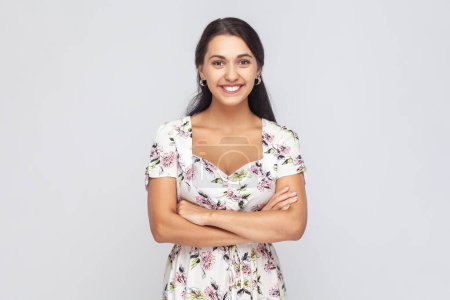 Foto de Portrait of satisfied attractive woman wearing white dress standing with crossed arms and smiling happily, looking at camera. Indoor studio shot isolated on gray background. - Imagen libre de derechos